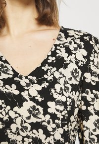 Kvinna iklädd en svart klänning med stora vita blommönster och en V-ringning som visar nyckelbenet och en del av axellängden på sitt bruna hår.