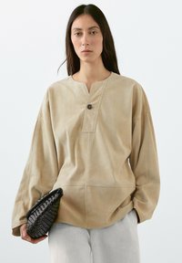 Beige Lederpullover mit Knopfleiste, weiten Ärmeln und Nahtdetails. Kombiniert mit einer schwarzen gewebten Clutch.