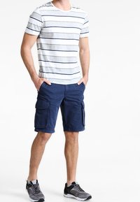 Muž v bílošedivém pruhovaném tričku, tmavě modrých cargo šortkách s kapsami a šedočerných teniskách, stojí s rukama v kapsách.