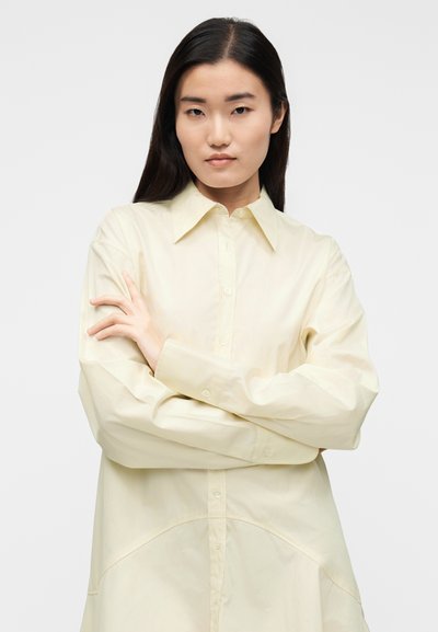 Šviesiai geltona marškinėliai su sagomis, klasikiniais apykakle, ilgomis rankovėmis ir lenktu apačia. Lygus audinys su laisvu prigludimu.