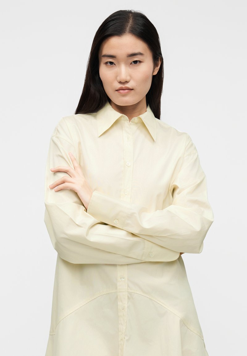 Chemise boutonnée jaune pâle avec un col classique, des manches longues et un ourlet courbé. Tissu lisse avec une coupe décontractée.