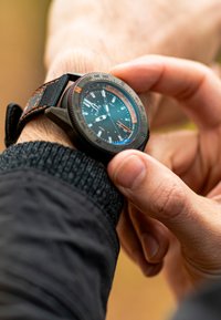 Montre-bracelet noire et orange avec une lunette en métal et un bracelet texturé, arborant un cadran sombre avec des index et des aiguilles horaires blancs et colorés.
