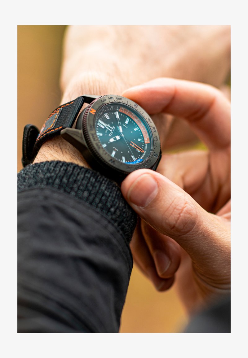 Sort og orange armbåndsur med metal bezel og struktureret rem, prydet med en mørk urskive med hvide og farverige timemarkører og visere.