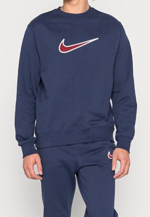 Mężczyzna w granatowej bluzie i pasujących spodniach, oba z czerwono-białym logiem Nike swoosh.