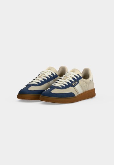 Ζεύγος sneakers χαμηλού τύπου με μπεζ και ναυτικό πάνελ, λευκά κορδόνια, καφέ σόλες και μικρό λογότυπο Polo στη πλευρά.