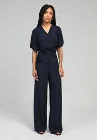 Combinaison en bleu marine avec manches bouffantes, design à jambes larges et ceinture. Fabriquée en tissu lisse, avec un décolleté en v et une coupe ajustée.