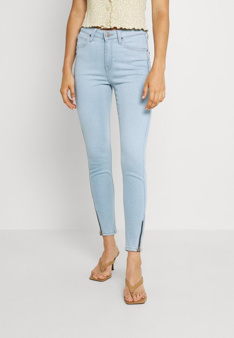 Jeans skinny de color azul claro con detalle de cremallera en el tobillo, hechos de un material de denim elástico, combinados con una blusa corta con estampado.