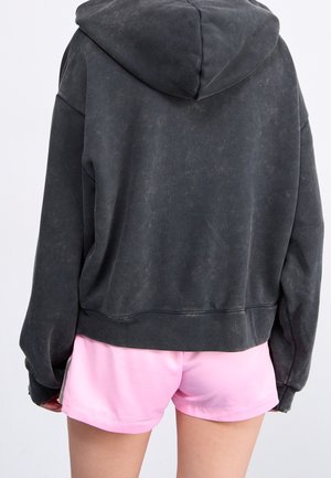 Personne portant un sweat à capuche noir délavé et un short rose vif, vue de dos sur un fond clair uni.