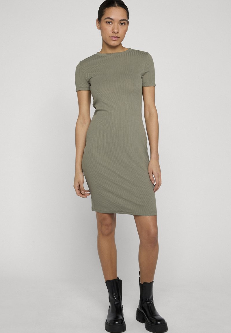Femme debout portant une robe côtelée olive courte à manches courtes, longueur au genou, et des bottines noires épaisses, sur fond clair uni.