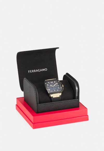 Hodinky FERRAGAMO s obdlžnikovým čiernym ciferníkom, zlatými akcentmi, modrým číselníkom a gumovým remienkom, prezentované v čiernej a červenej krabičke.