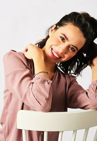 Blusa rosa chiaro, texturizzata, con maniche lunghe e dettagli con bottoni, indossata da una donna seduta su una sedia bianca, sorridente con capelli scuri e disordinati.