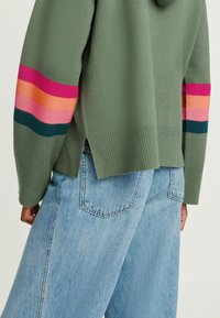 Felpa verde a maglia con maniche a righe rosa, arancioni e teal, con orlo a costine e spacchi laterali, abbinata a jeans azzurri chiari.