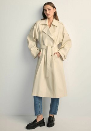 Kobieta ubrana w beżowy płaszcz trencz z paskiem, nałożony na niebieskie dżinsy i czarne mokasyny, stoi z rękami w kieszeniach płaszcza na jednolitym tle.