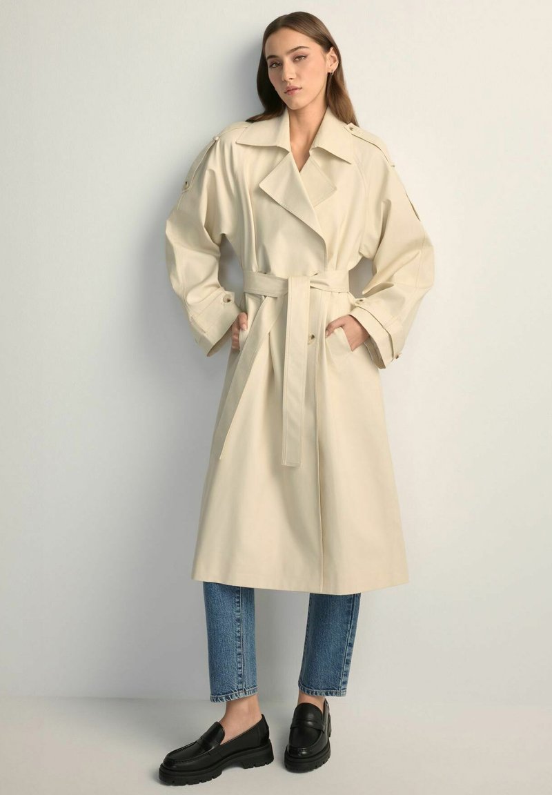 Kobieta ubrana w beżowy płaszcz trencz z paskiem, nałożony na niebieskie dżinsy i czarne mokasyny, stoi z rękami w kieszeniach płaszcza na jednolitym tle.