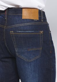 Donkerblauwe jeans van denim met een leren patch, oranje stiksels en een vijf-pocket ontwerp. Bevat subtiele vervaging op de achterzak.