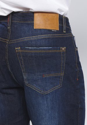 Mørkeblå denimjeans med et læderpatch, orange syninger og fem-lomme design. Har subtilt slid på baglommen.
