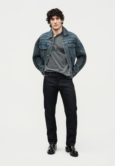 Νεαρός με σγουρά μαλλιά που φοράει μια ξεθωριασμένη μπλε τζιν ζακέτα πάνω από μία γκρι φθαρμένη μπλούζα, μαύρο τζιν και μαύρα γυαλιστερά μποτάκια, στέκεται όρθιος.