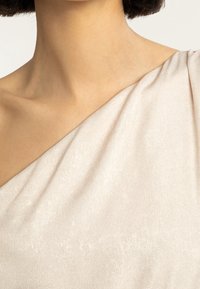 Sukienka na jedno ramię z miękkiego, błyszczącego beżowego materiału. Gładka faktura elegancko się układa, lekko połyskując i odsłaniając skórę na ramieniu.