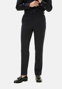 Pantaloni blu scuro con una vestibilità su misura, frontale plissettato e passanti per cintura, realizzati in un tessuto liscio. Abbinati a mocassini neri.