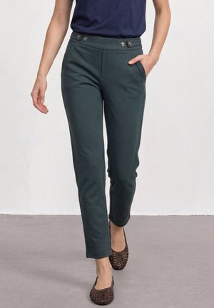 Femme portant un pantalon slim vert foncé avec des boutons à la taille et des chaussures plates tressées marron, debout contre un mur uni.