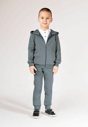 Szara bluza rozpinana na zamek z kapturem, w zestawie z dopasowanymi spodniami. Bluza ma dwie przednie kieszenie i ściągacze na rękawach. Czarne trampki z białą podeszwą.