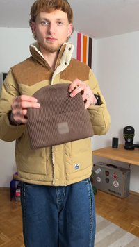 Bruine gebreide beanie met ribbelstructuur, voorzien van een omgeslagen boord en een klein logopatch; vastgehouden door een persoon in een beige cordjacket.