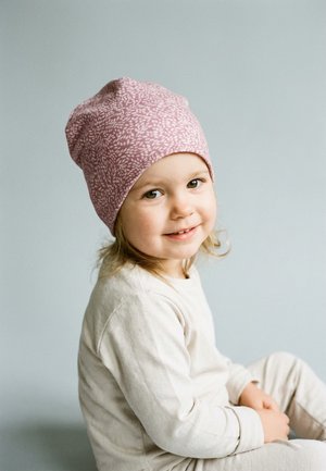 Lächendes Kleinkind mit hellbraunen Haaren, das eine rosa gemusterte Mütze und ein cremefarbenes Langarmshirt trägt und vor einem hellgrauen Hintergrund sitzt.