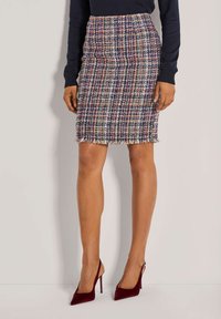 Tweed potloodrok in meerkleurige ruit met franjes aan de onderkant. Gecombineerd met bordeauxrode hoge hakken. De textuur lijkt ruw en geweven.