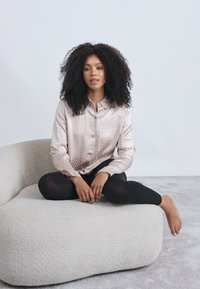 Ανοιχτό ροζ σατέν πουκάμισο με κουμπιά και υφή, συνδυασμένο με μαύρους κολάν. Μοντέλο καθισμένο σε ένα μαλακό, καμπυλωτό, λευκό μαξιλάρι.