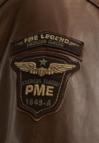 Smeđa kožna etiketa s izvezenim tekstom: "PME LEGEND AMERICAN CLASSIC," uz ukrasna krila i zvijezdu. Šivana detalja oko rubova.
