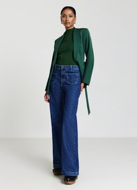 Blazer lungo a maniche lunghe di colore verde strutturato sopra una maglia a coste verde scuro, abbinato a jeans in denim blu a vita alta e gamba larga, e scarpe con platform nere.