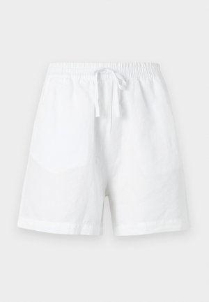 Nő áll, fekete ujjatlan felsőt, laza fehér gombos inget, fehér zsinóros rövidnadrágot és bézs hegyes orrú, bokapántos lapos cipőt visel.