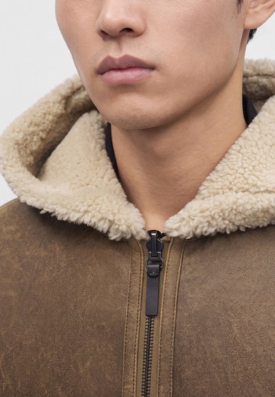 Close-up of a person wearing a brown shearling jacket with a beige fluffy collar and a black zipper pull tab.