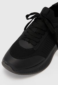 Chaussure de sport noire avec une tige en mesh respirant, motif texturé et lacets plats ; semelle en caoutchouc avec une finition lisse et un design de bout arrondi.