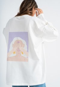 Witte oversized sweatshirt met een kleurrijke illustratie van een persoon met lang haar dat hun ogen bedekt, tegen een lichtpaarse achtergrond.