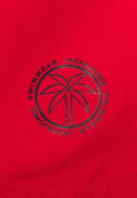 Rode stoffen badkleding met een cirkelvormig zwart logo waarop een palmboom staat en de tekst "JACK & JONES" en "SWIMWEAR" rondom het ontwerp.