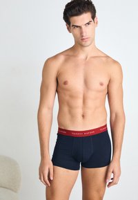 Sötét tengerészkék férfi boxer alsónadrág piros derékrésszel, melyen a 'TOMMY HILFIGER' logó látható. Simaság, testhezálló forma és közepes magasságú dizájn jellemzi.
