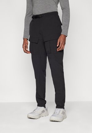 Personne portant un pantalon cargo noir avec des poches à rabat, un t-shirt gris à manches longues et des baskets blanches épaisses, debout devant un fond uni.