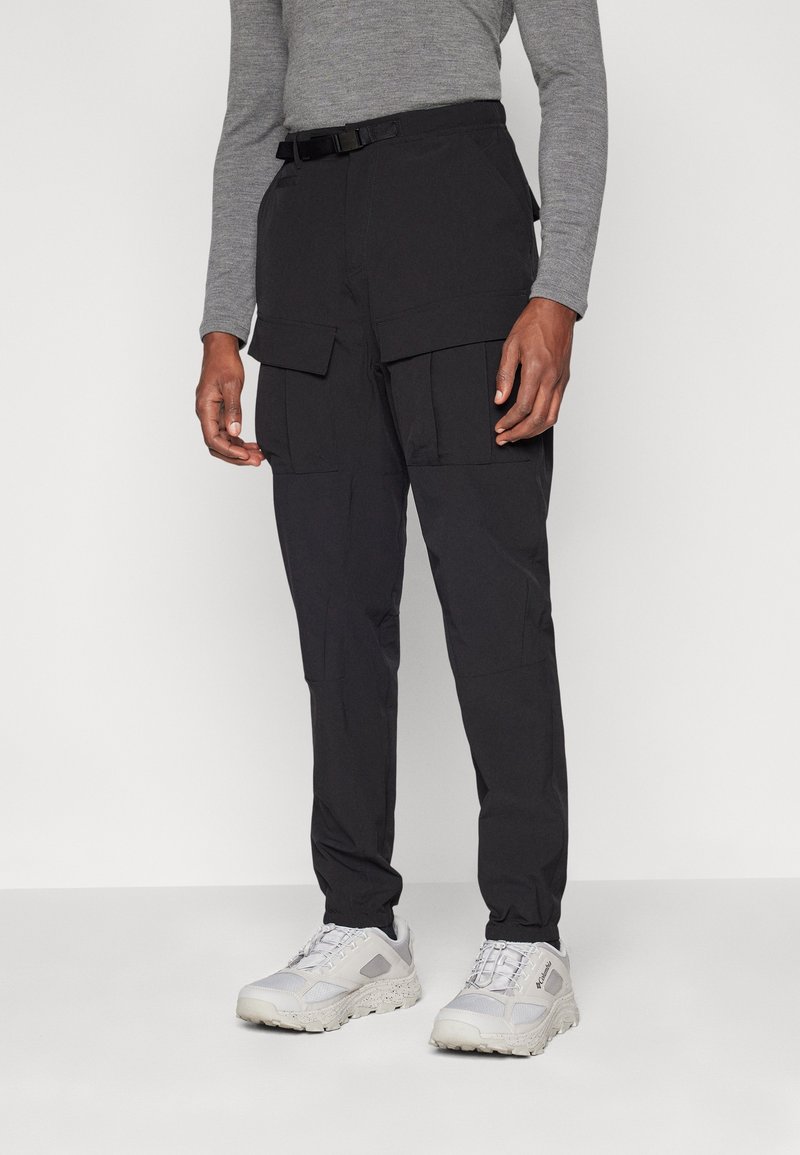 Personne portant un pantalon cargo noir avec des poches à rabat, un t-shirt gris à manches longues et des baskets blanches épaisses, debout devant un fond uni.