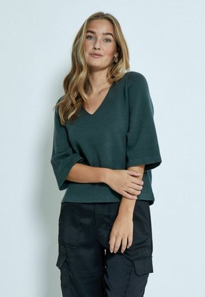 Pull vert, à col en V, à manches courtes, fait de matière douce, avec une coupe décontractée et un design minimaliste, associé à un pantalon cargo noir.