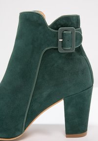 Bottine en daim vert foncé à talon bloc avec une bride à boucle verte assortie près du bord supérieur.