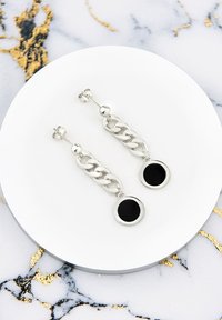 Silverkedjear med cirkulära svarta accenter. Har en slät yta och en hängande design, visad på en vit tallrik mot en marmorerad bakgrund.