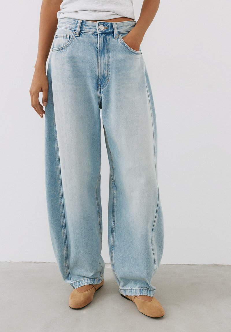 Personne portant un jean en denim bleu clair ample, un haut blanc et des chaussures plates beige, debout avec une main dans la poche sur un sol gris.
