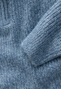 Gaismas zila adīta džemperis ar teksturētu ribu dizainu un salocītu piedurkni, izceļot mīkstu, šķiedraina materiāla struktūru.