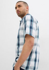 Camicia a maniche corte a quadri nei toni del bianco, blu e grigio. Presenta un colletto con bottoni e una vestibilità rilassata, con una texture simile al cotone.