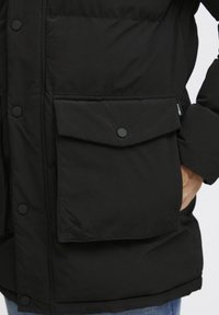 Chaqueta acolchada negra con cierre frontal de botones, gran bolsillo de solapa y textura suave. El material parece ser resistente al agua y duradero.