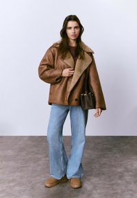 Chaqueta de piel marrón con solapas anchas, combinada con vaqueros azules acampanados y botas tobilleras color canela. Un pequeño bolso marrón realza el conjunto.