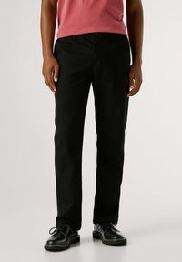 Zwarte broek van gladde stof, recht model, gecombineerd met een rood shirt en zwarte schoenen met ronde neus, voorzien van vetersluiting.