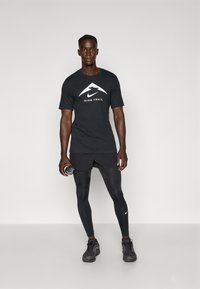 Schwarzes Nike Trail-T-Shirt mit weißem Logo, kombiniert mit schwarzen Shorts und enganliegenden Leggings. Das Model hält eine Wasserflasche und trägt dunkle Sportschuhe.