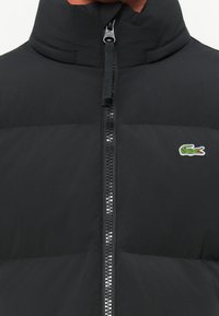 Doudoune noire avec un col haut, une fermeture éclair à l'avant et un petit logo crocodile vert. Présente un tissu texturé avec des coutures horizontales.